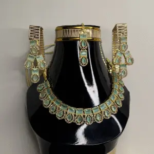 A gold necklace set with matching earrings and a choker, featuring green gemstones, displayed on a black mannequin bust.